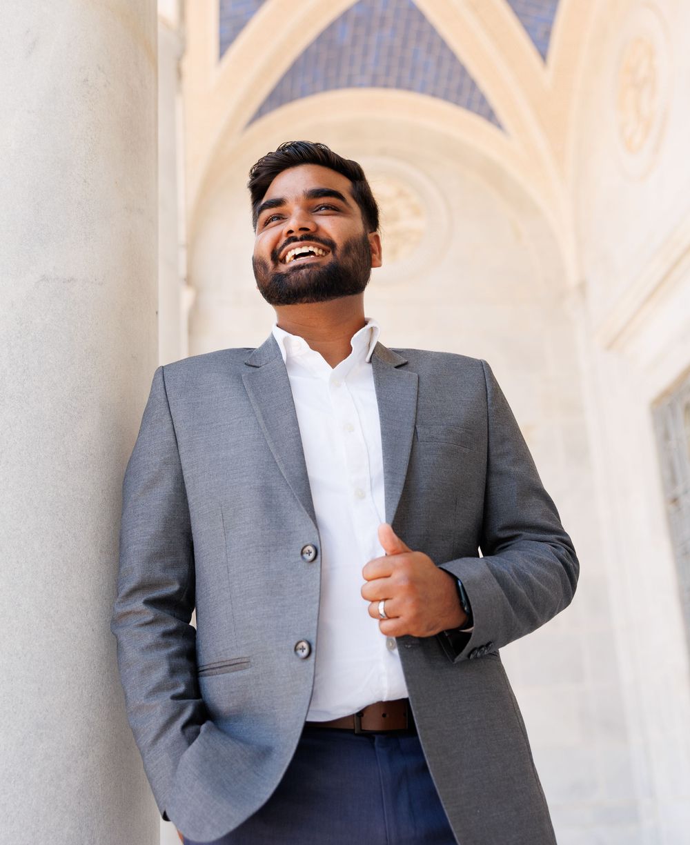 Smiling young man in gray blazer and white shirt laughing outdoors near ornate stone archway columns.