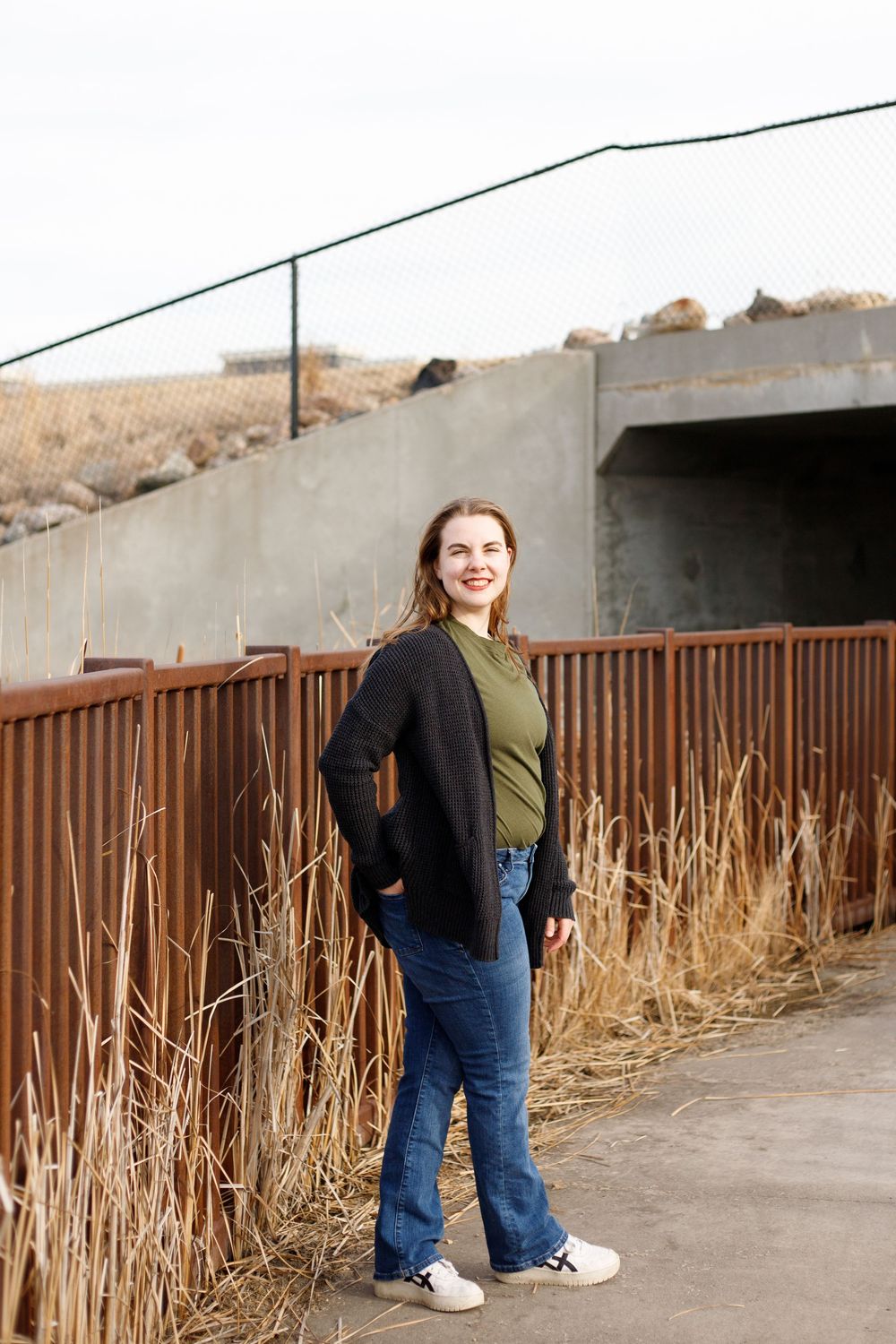 Young woman in black cardigan, green top, and jeans smiling outdoors near rustic fence and dry grass.