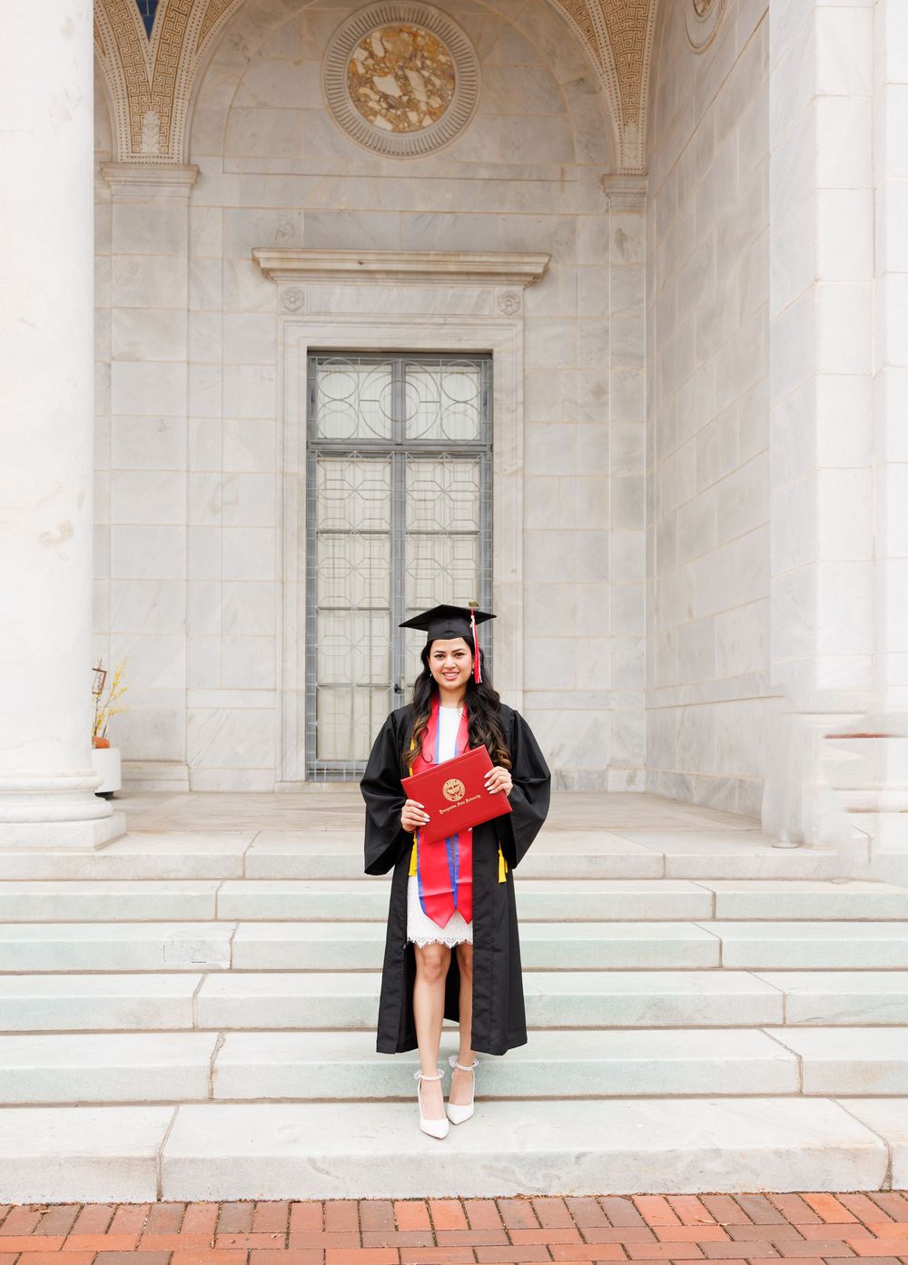 Graduate in black cap and gown holding red diploma folder on marble steps of a university building.