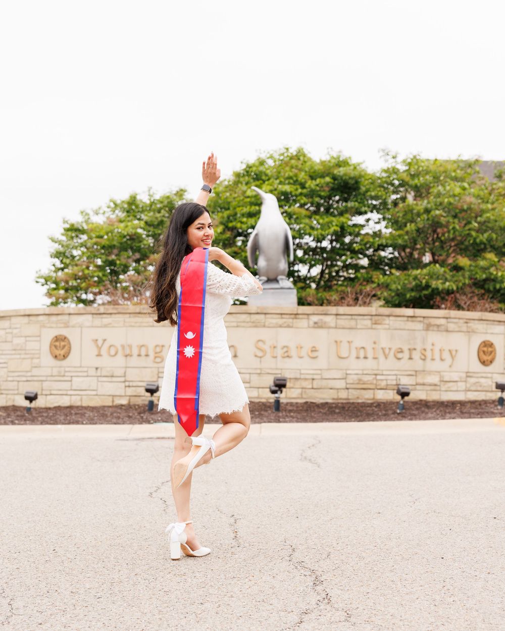 Young woman in white dress with red graduation stole poses joyfully in front of Youngstown State University sign.