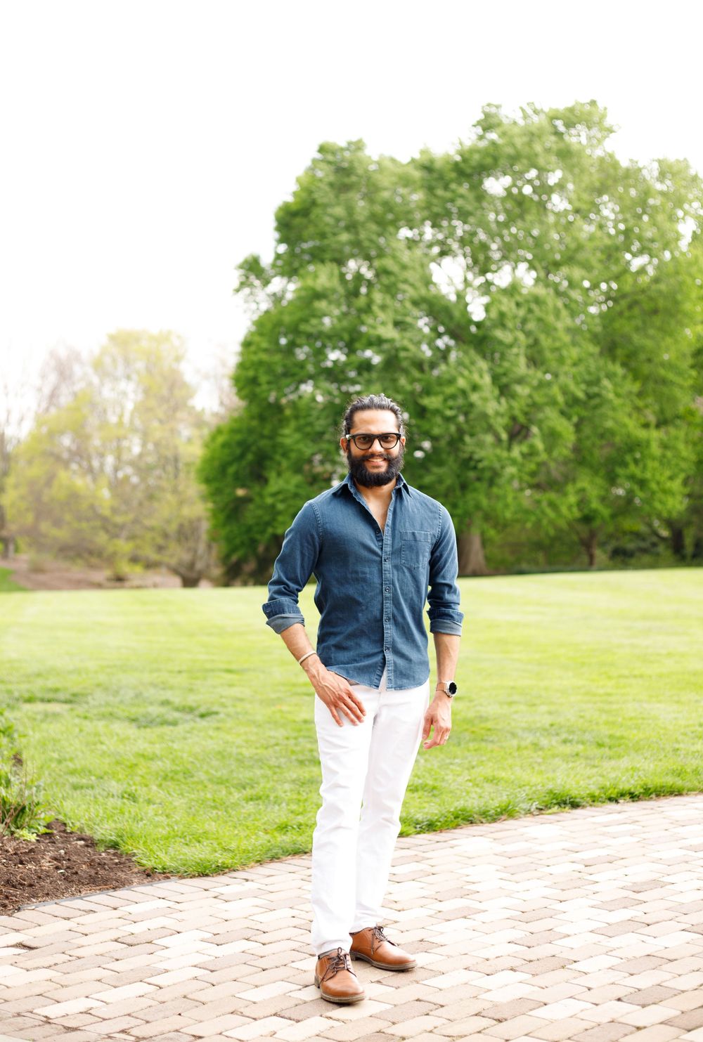 Man in blue shirt and white pants smiling outdoors on brick path with lush green trees in background.