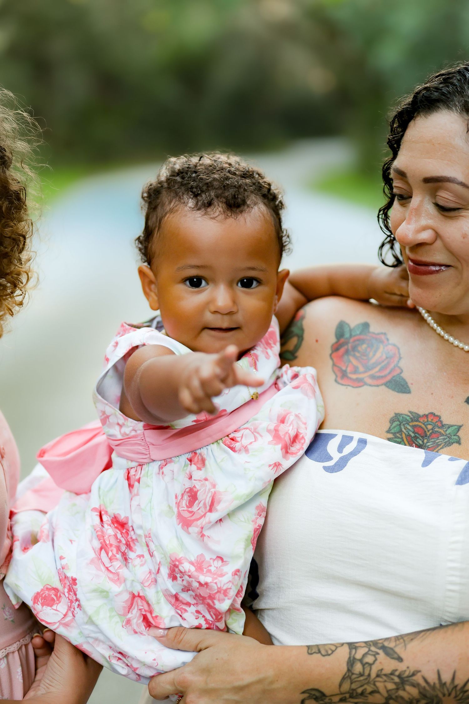 A tattooed woman holds a smiling baby girl in a floral dress outdoors, as the baby reaches toward the camera.