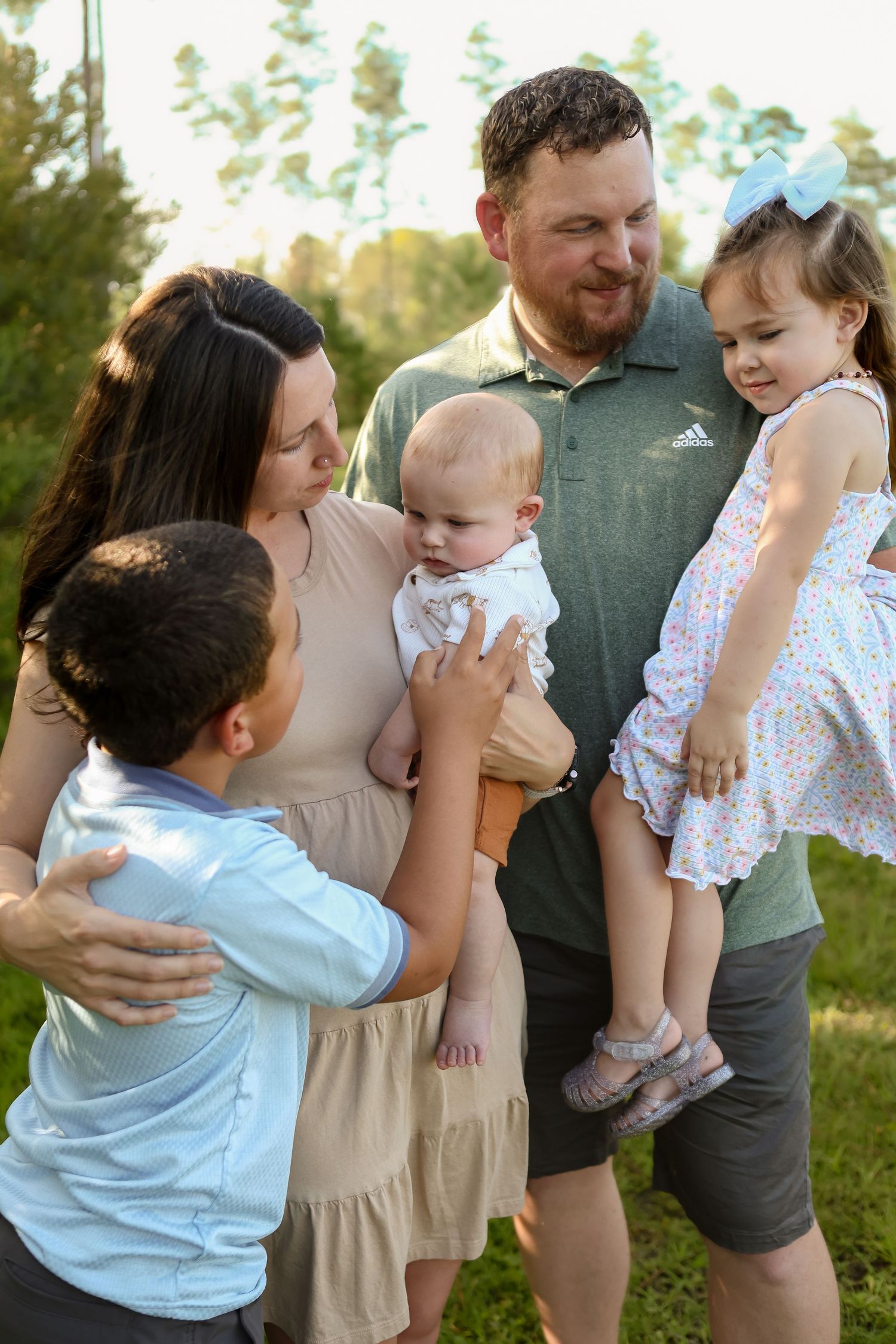 A happy family of five poses outdoors, parents holding three young children, smiling together on a green lawn.