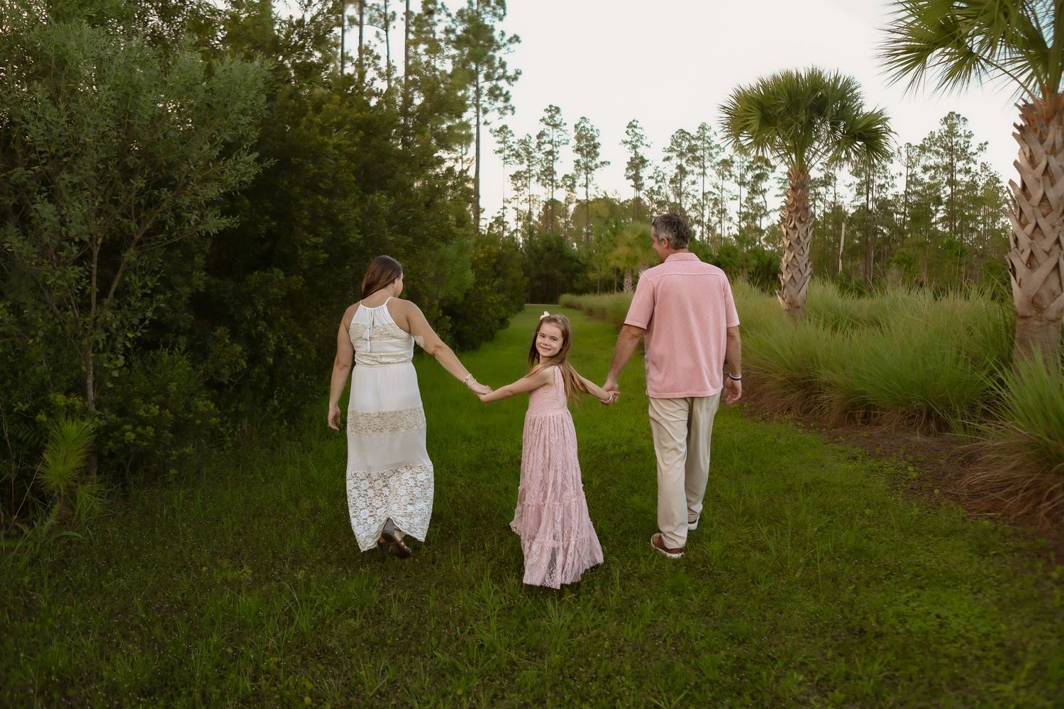 Family of three holding hands walking through lush green park, parents in white and pink outfits with young girl in pink dress.