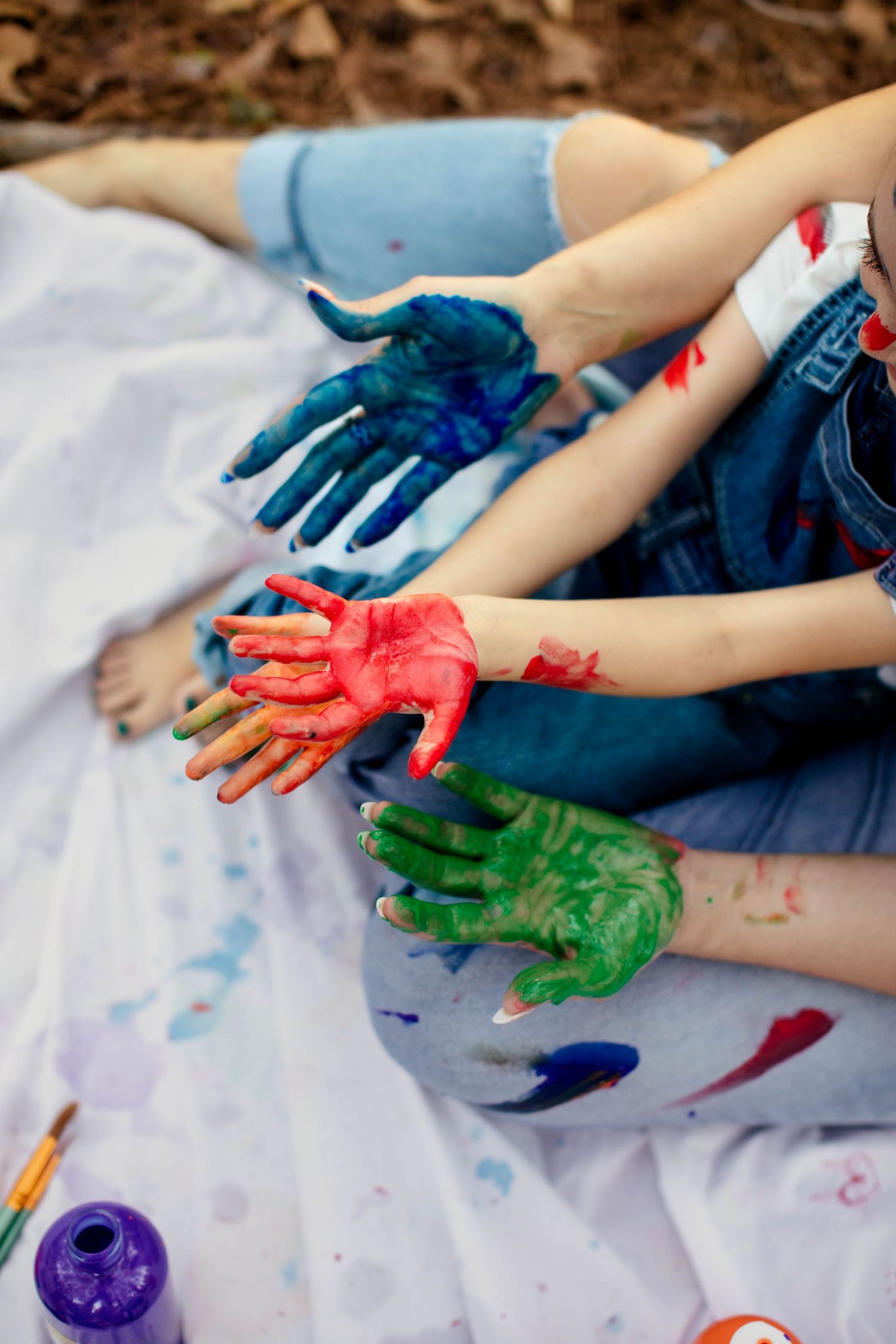 Paint-covered hands in blue, red and green finger paint sitting on a white blanket outdoors.