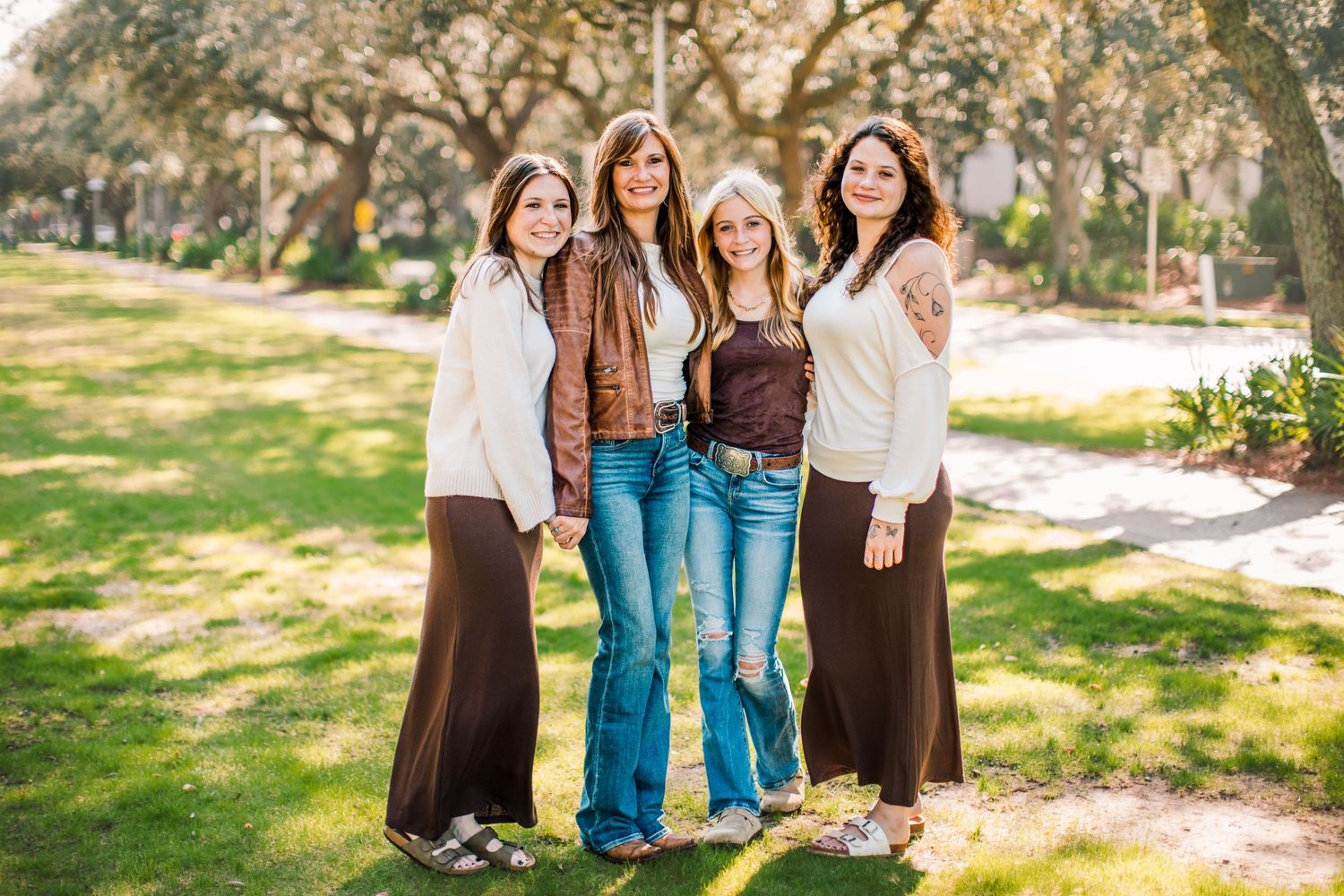 Four friends pose together outdoors in casual clothing on a sunny day with trees and grass in the background.