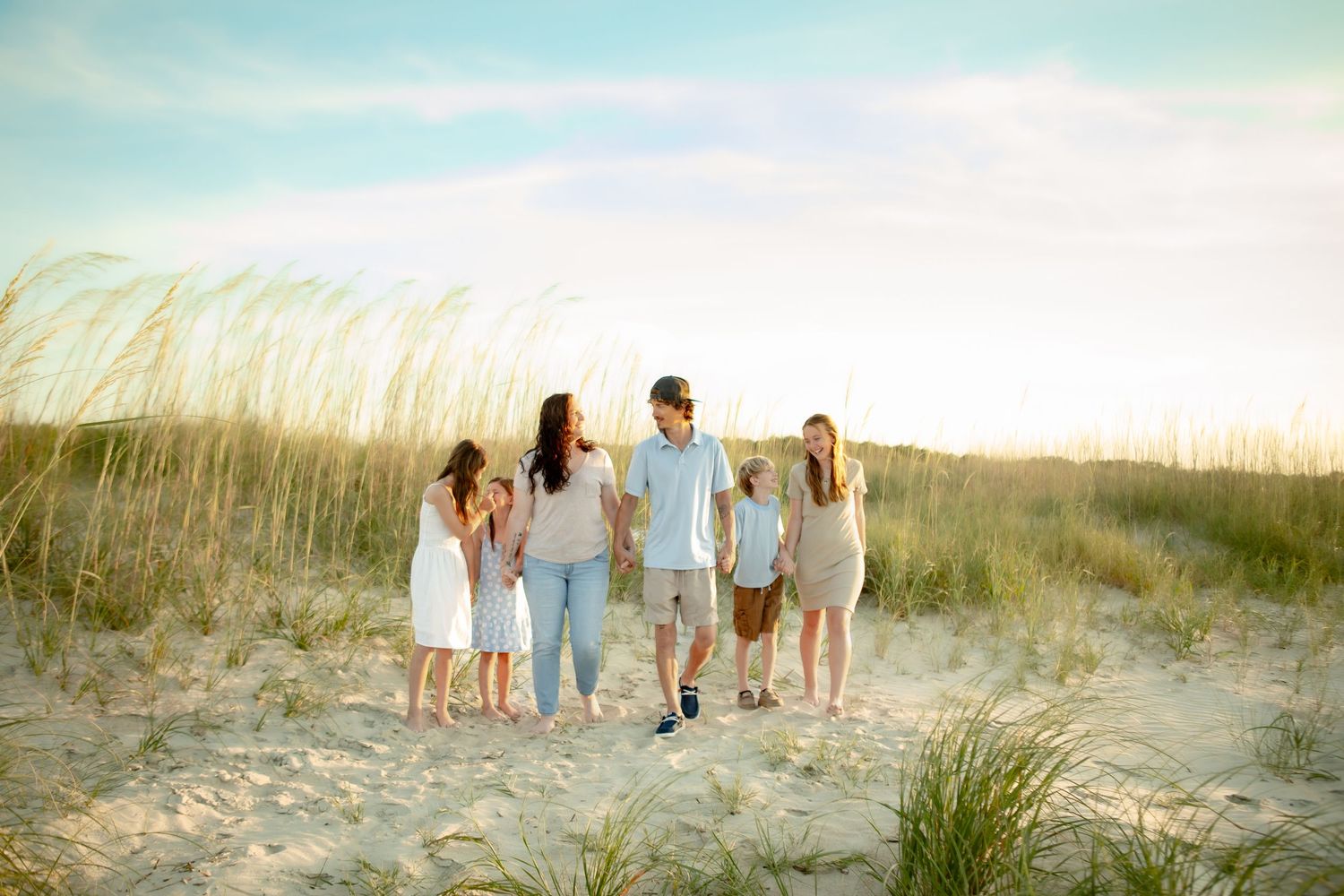 Family of six walking together on sandy beach dunes with sea grass and blue sky backdrop.
