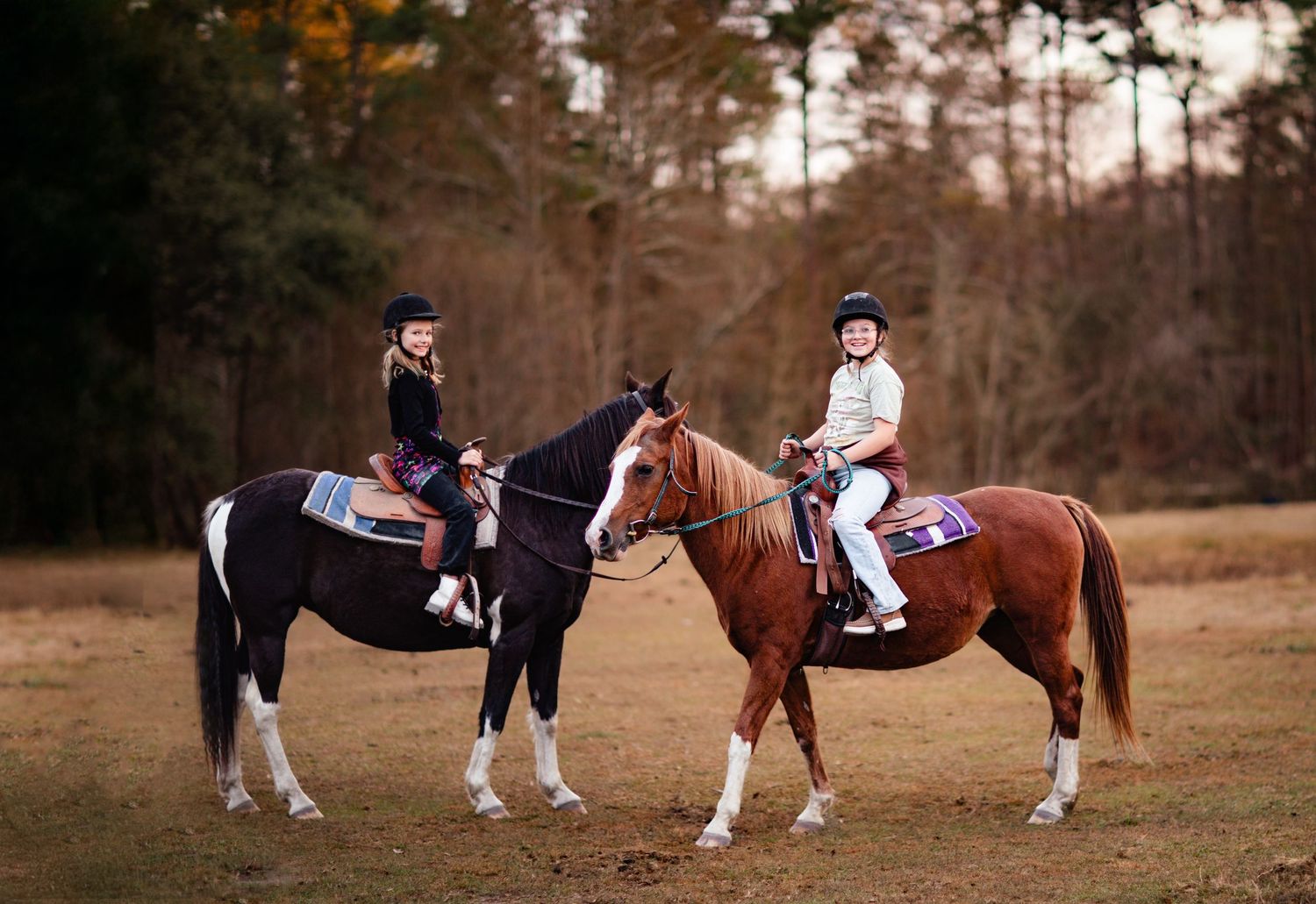 Two riders on horseback in equestrian attire sit atop their horses in a rustic outdoor setting during autumn.