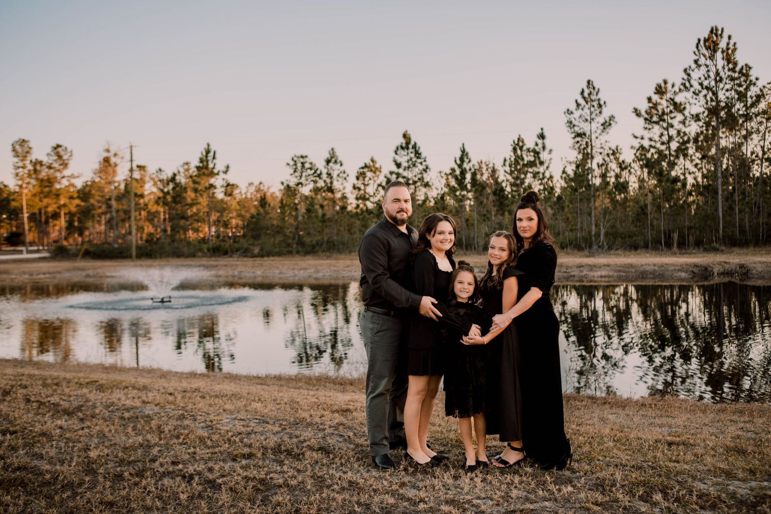 Group wearing black and dark outfits poses together near reflective pond at sunset with pine trees in background.
