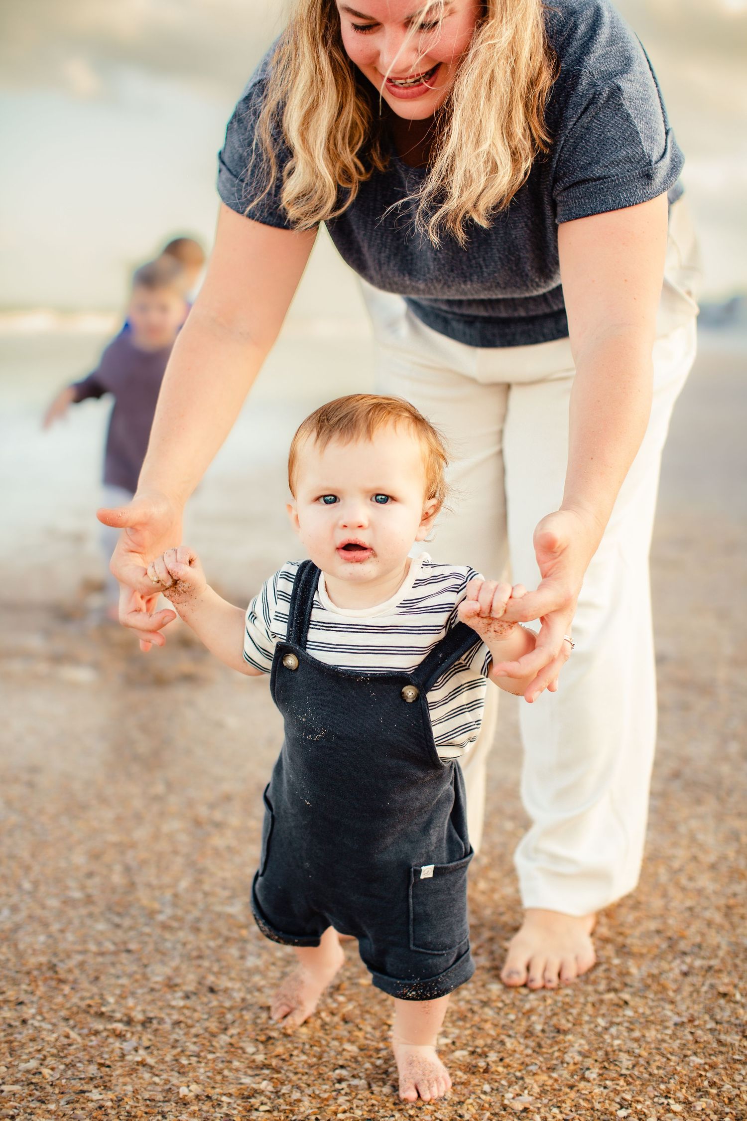 A parent helps a toddler in navy overalls take barefoot steps on a beach during a joyful moment together.