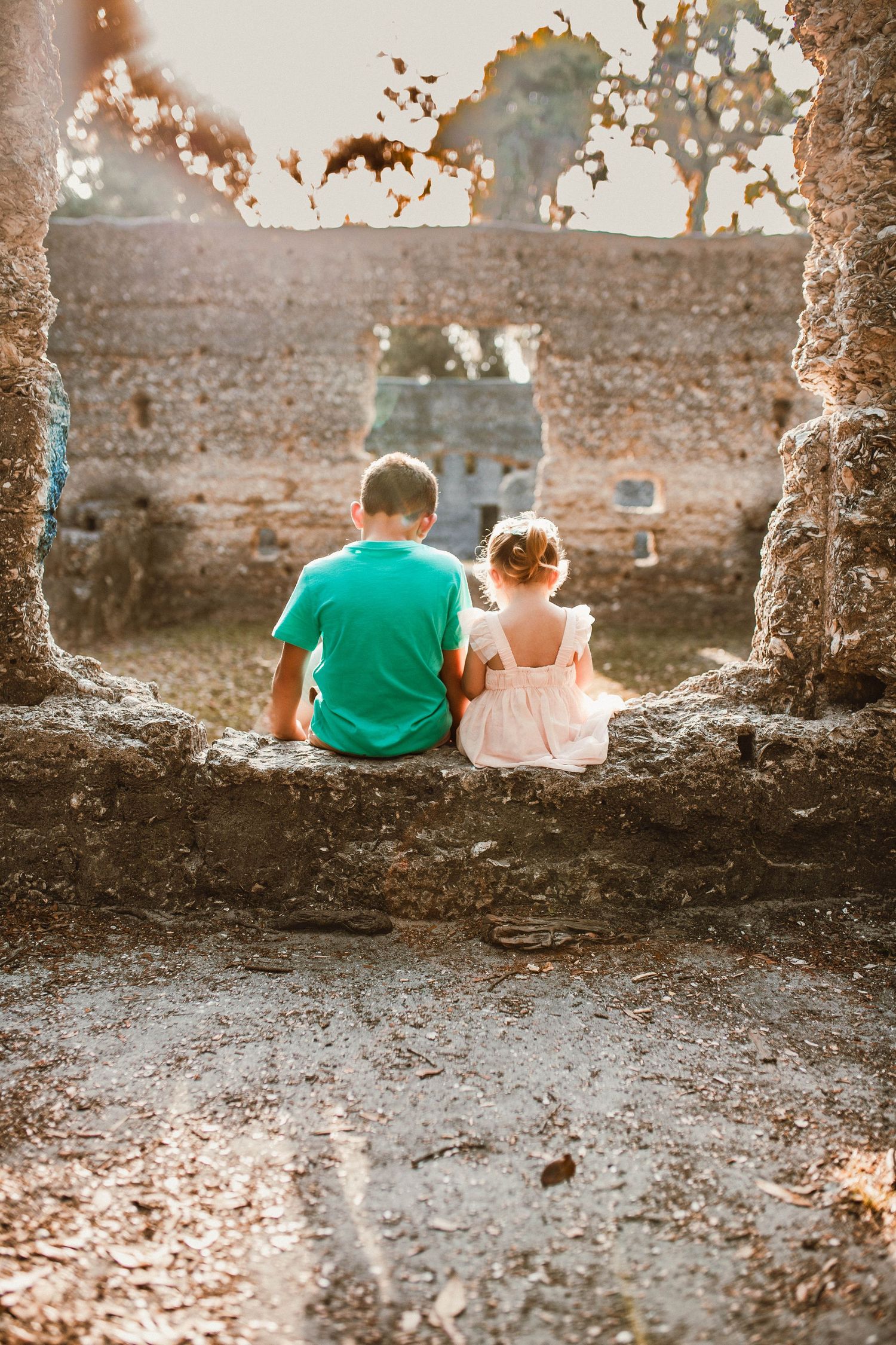 Two children sit together on a stone ledge at sunset surrounded by old stone ruins in warm golden light.