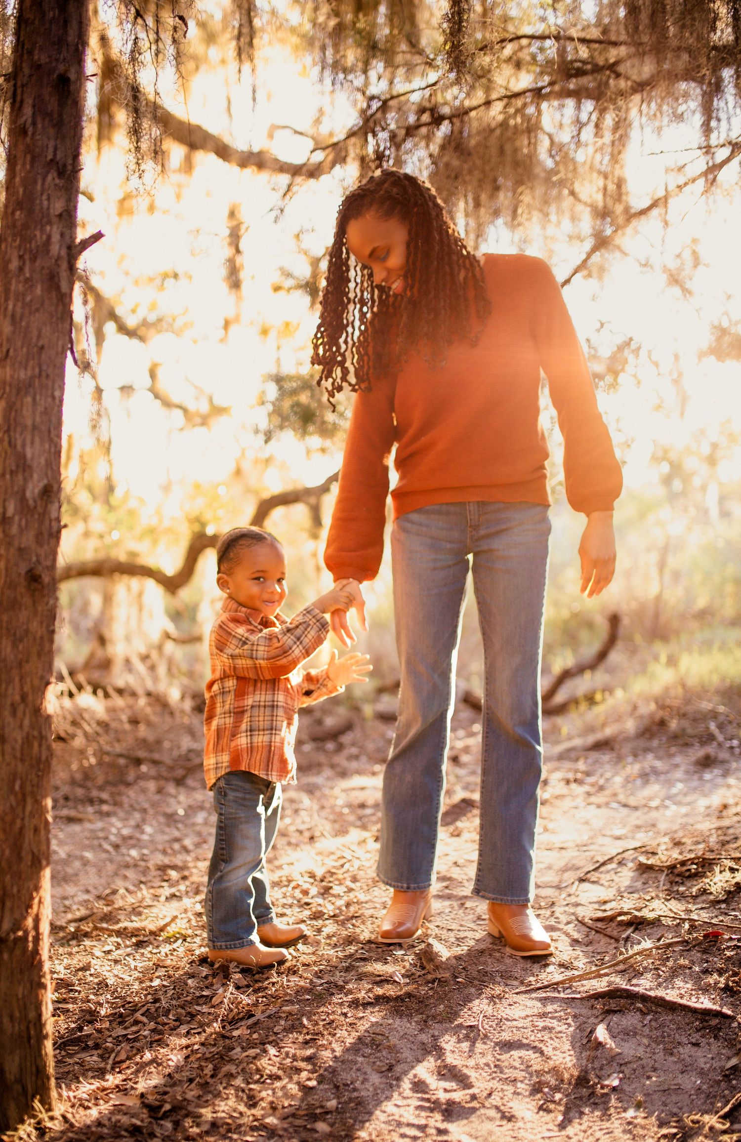 A small child and adult walk through a sunlit wooded area with golden light filtering through the trees.