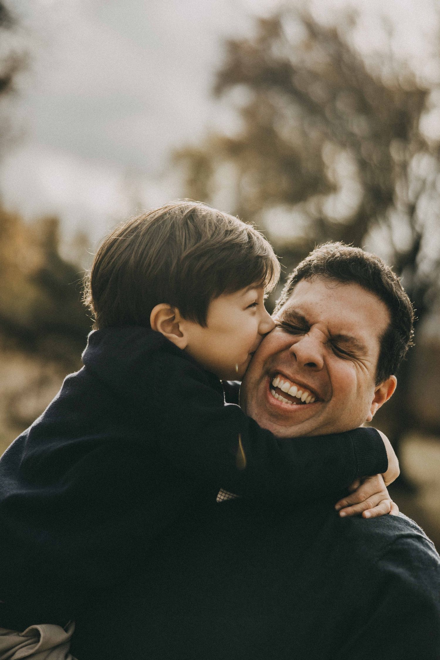 A joyful candid moment captured between parent and child sharing a sweet kiss and laugh outdoors at sunset.