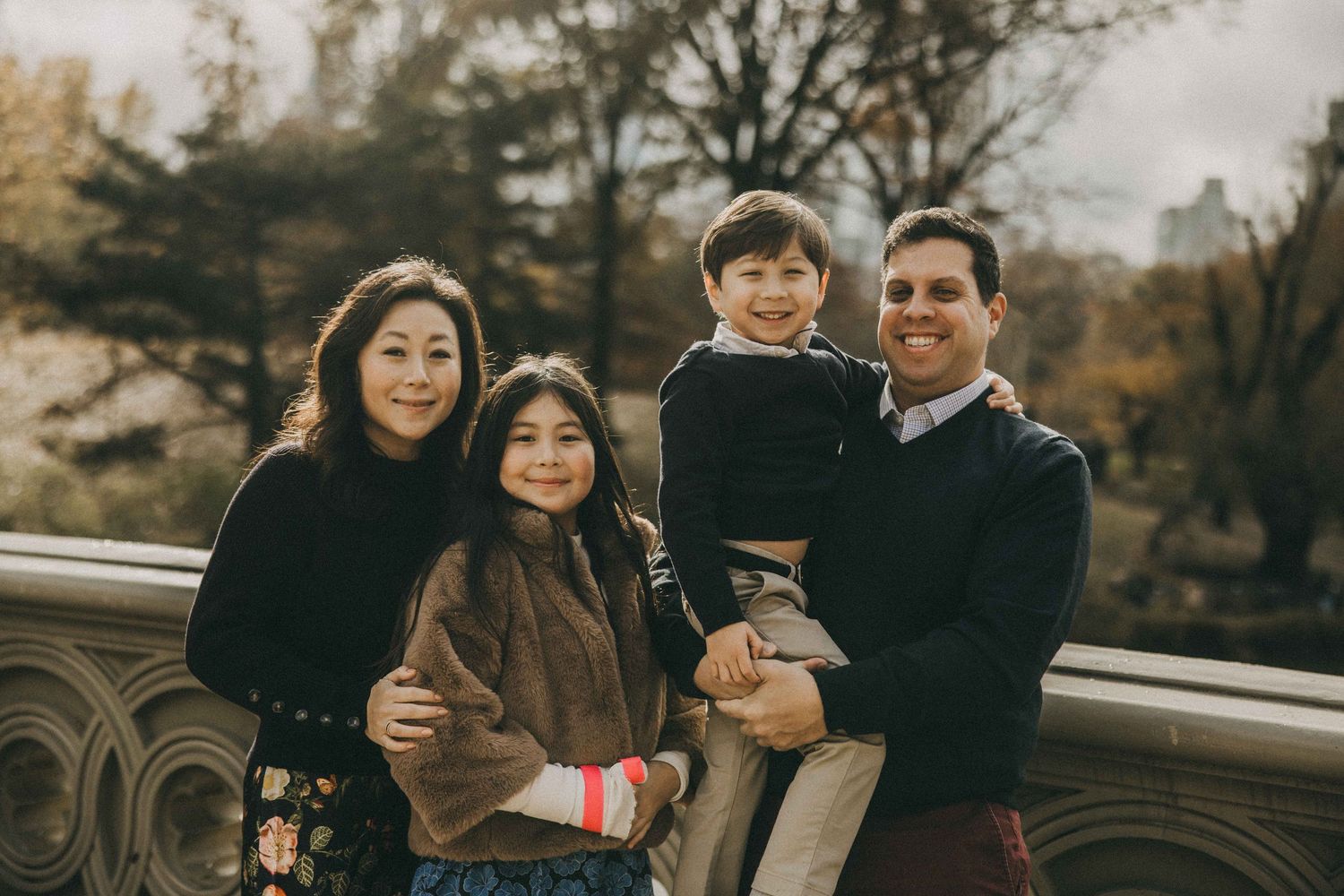A family stands together on a bridge in autumn wearing dark coats during a casual outdoor photoshoot.