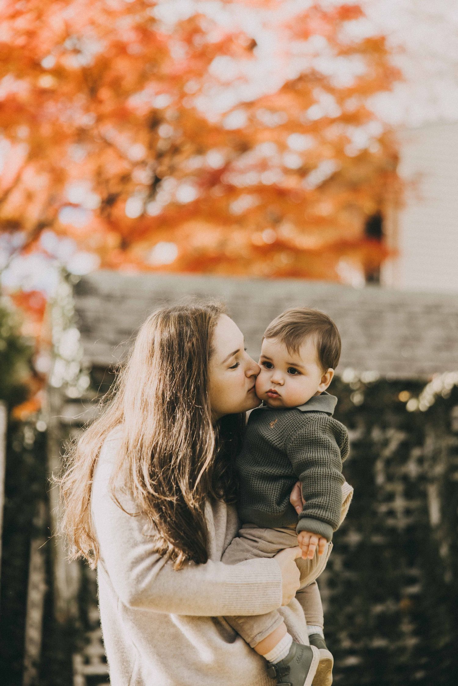 A parent and child share tender moments under vibrant orange autumn foliage against a stone wall backdrop.