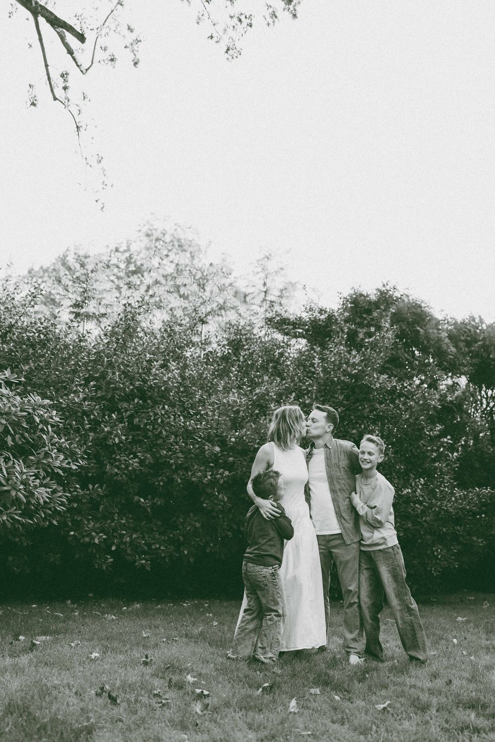 Black and white photo of a couple sharing a kiss outdoors while two children stand beside them in a lush garden.