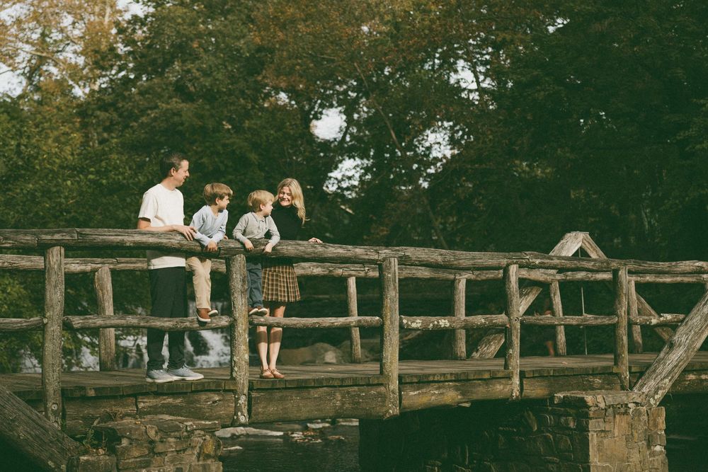 Family of four standing on a rustic wooden bridge surrounded by lush green trees in a park setting.