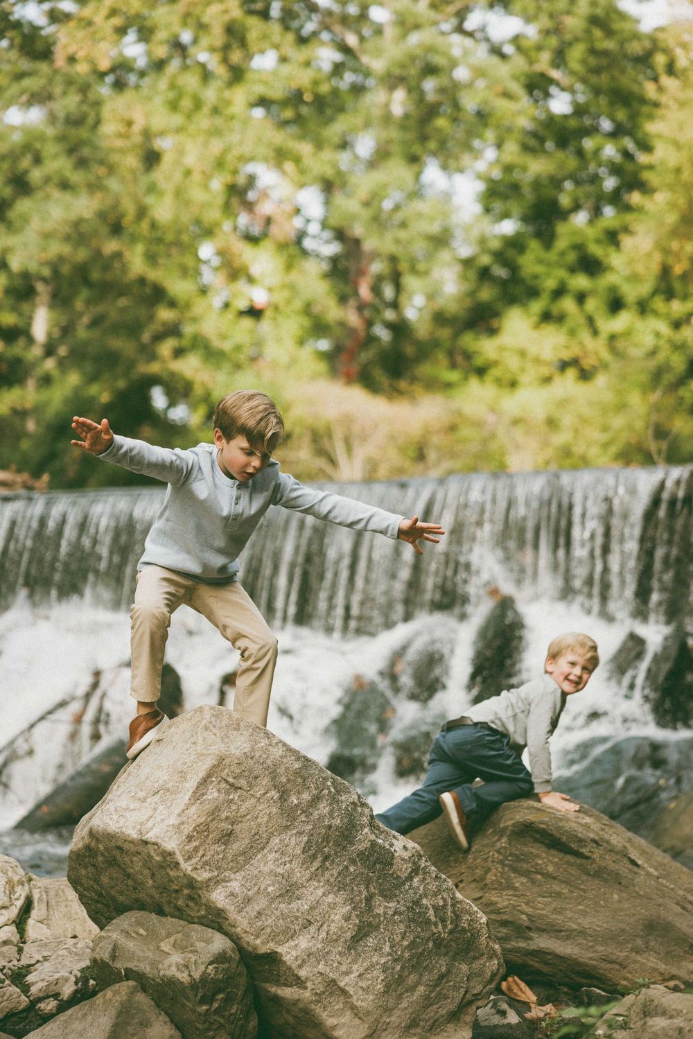 Two boys playing on rocks near a waterfall surrounded by lush green trees outdoors.