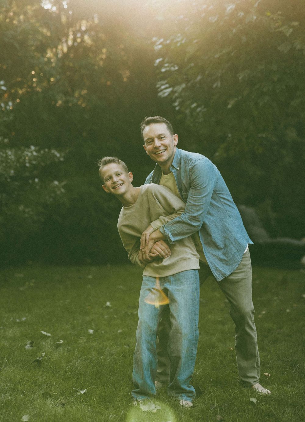 A smiling adult and child embrace playfully in a sunlit backyard surrounded by lush green trees.