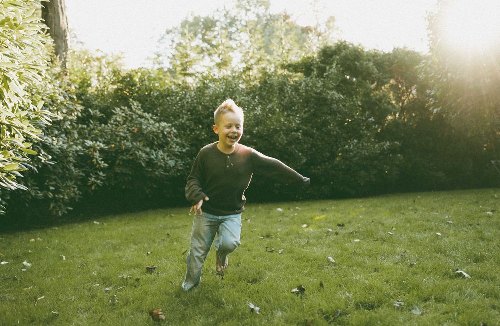 A joyful child running barefoot on green grass in a sunlit garden surrounded by lush bushes.