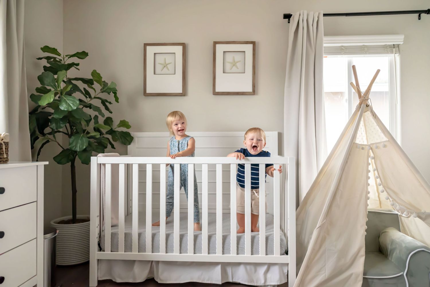 Toddler girl and her younger brother standing in a white crib laughing. In-home family session. Doughman Photography