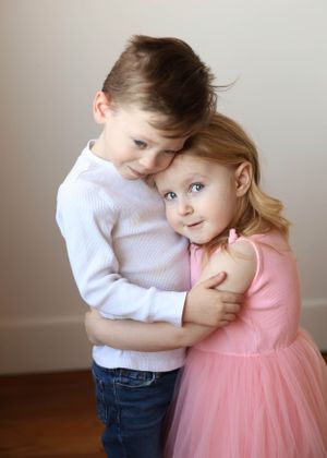 Two children share a sweet embrace, one wearing a white sweater and the other in a pink tulle dress.