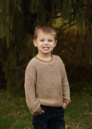 Child in brown sweater smiles warmly during outdoor fall photo session.