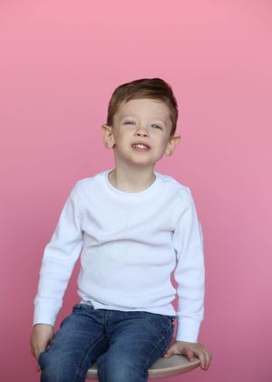 Child in white sweater and jeans poses playfully against pink backdrop.