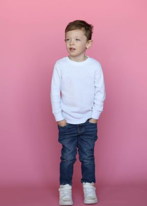 Child in casual white sweater and jeans stands against pink background.