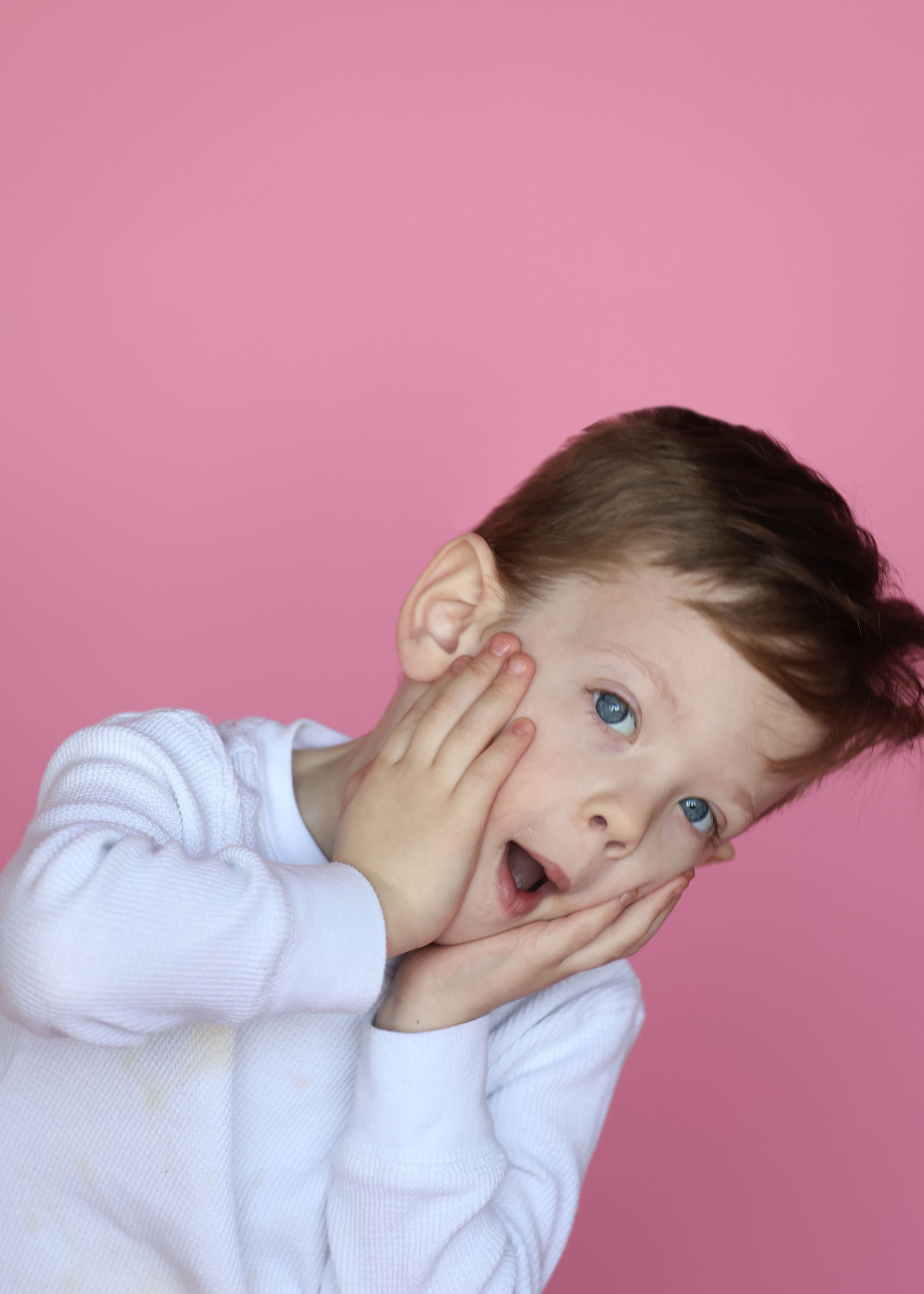 Child in white shirt makes playful expressions against pink background.