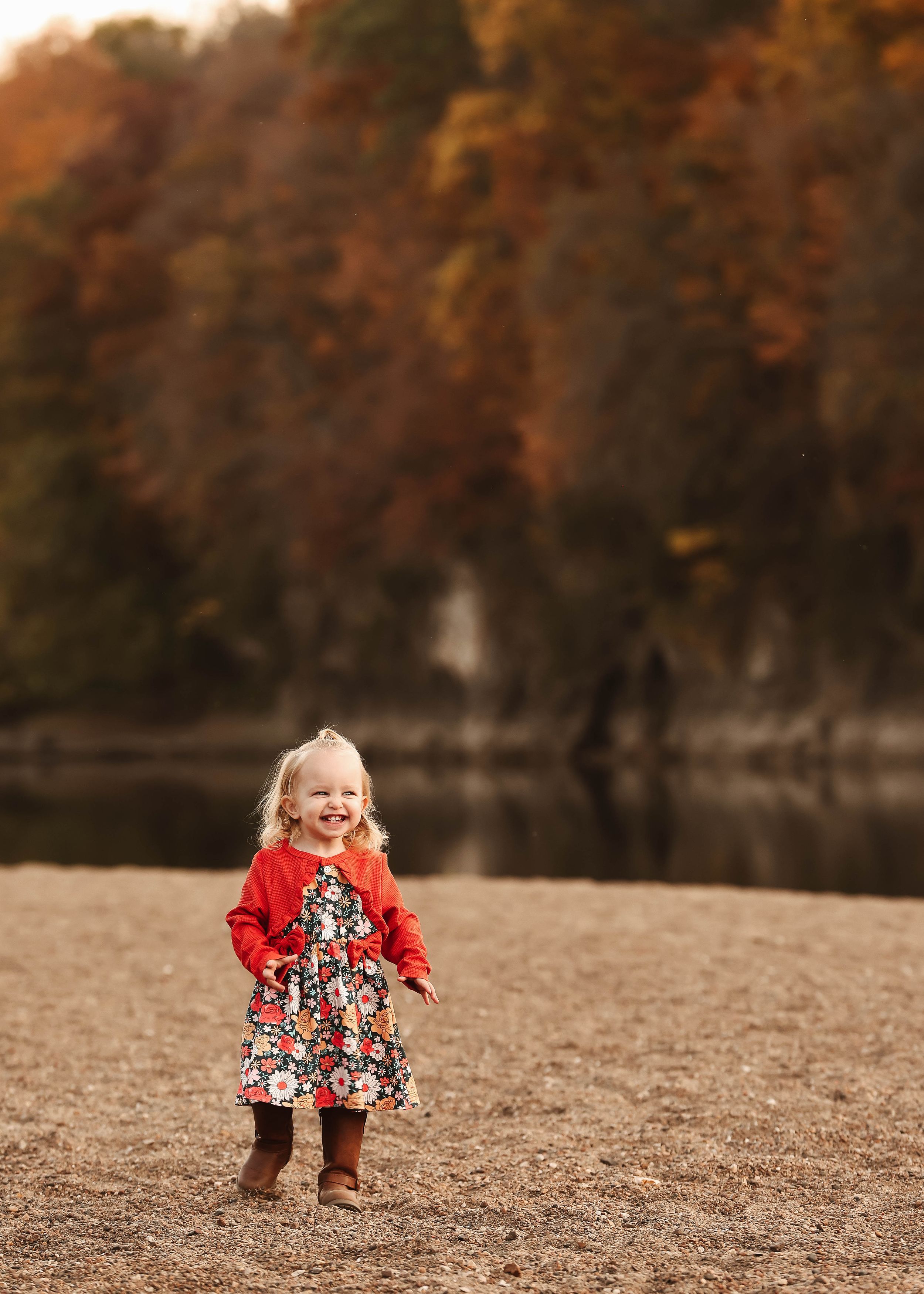 Child in red sweater playing on sandy beach with fall colors in background.