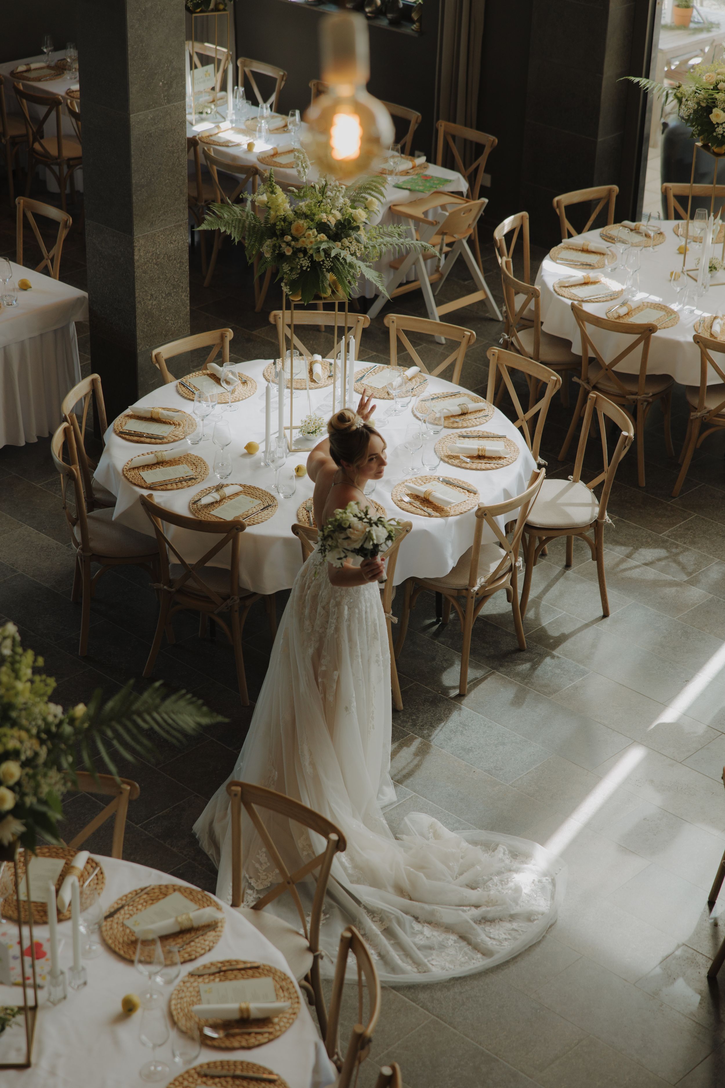 Elegant wedding reception setup with round tables, wooden cross-back chairs and flowing white decor in a rustic venue.