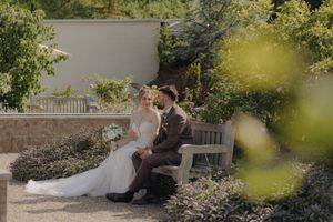 Two people in black and white wedding attire share an intimate moment seated on a wooden bench in a garden setting.