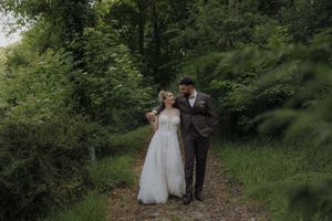 A couple in wedding attire walks together along a serene forest path surrounded by lush green foliage.