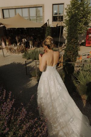 A figure in a flowing white strapless wedding dress walking outdoors near a garden with purple flowers.