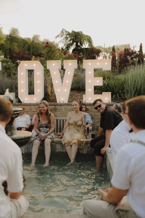 A group of wedding guests sits in front of illuminated LOVE letters at an outdoor evening celebration.