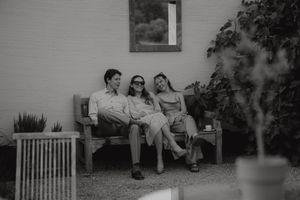 A black and white photo showing three people relaxing together on a wooden bench in a garden.