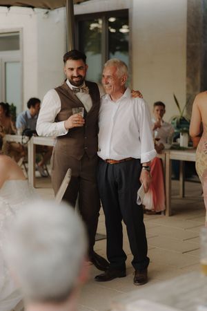 Two men sharing a moment at an indoor wedding reception with guests seated in the background.