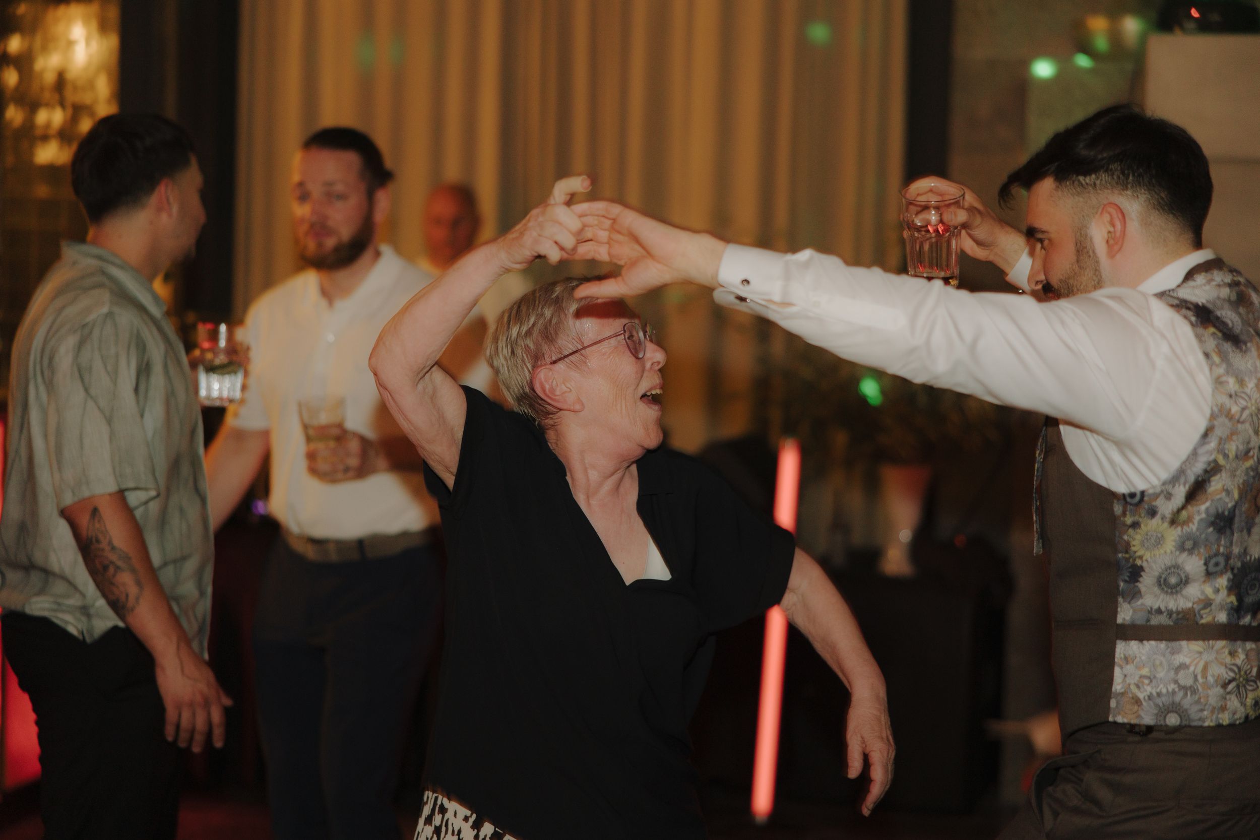 Happy dancing guests at a dimly lit wedding reception enjoy celebrating while holding drinks.