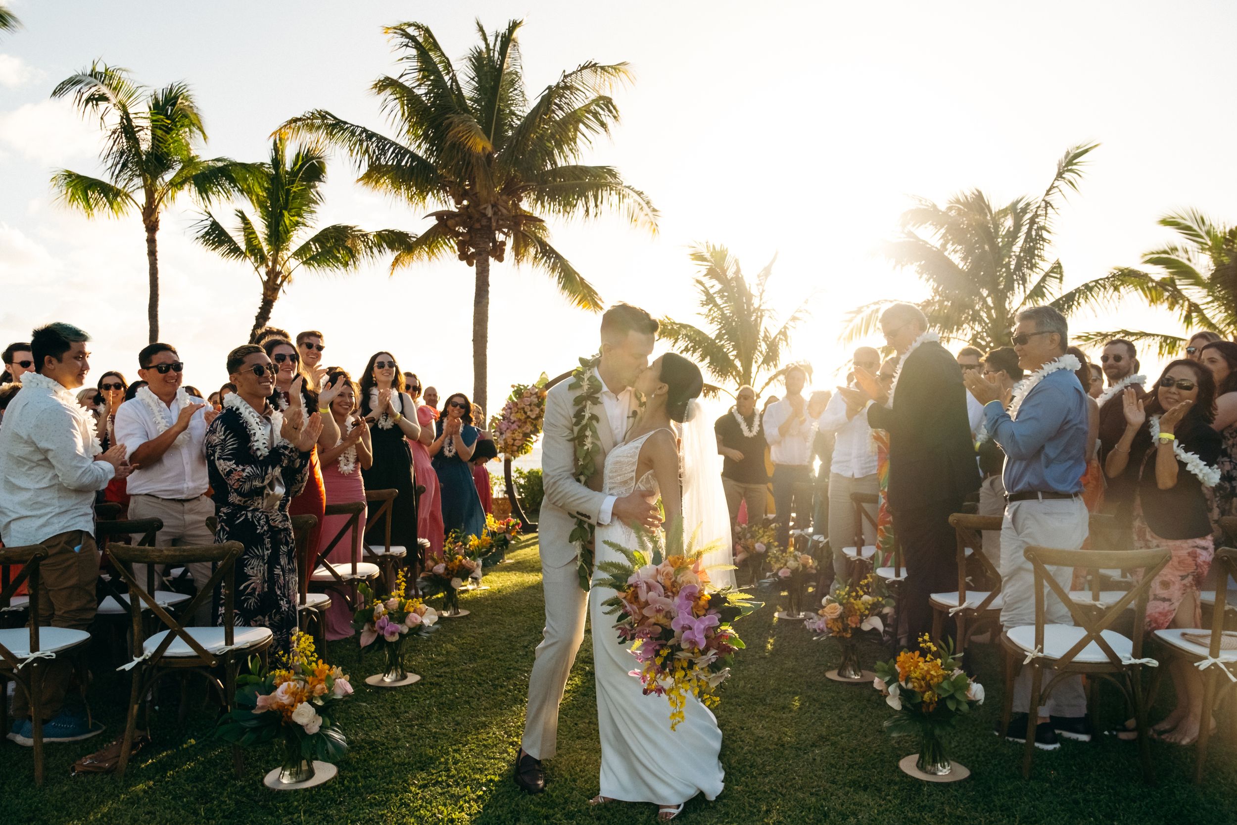 Bride and groom kissing after the wedding ceremony in the aisle, surrounded by guests at Lanikuhonua Venue, Ko Olina, Oahu, photographer