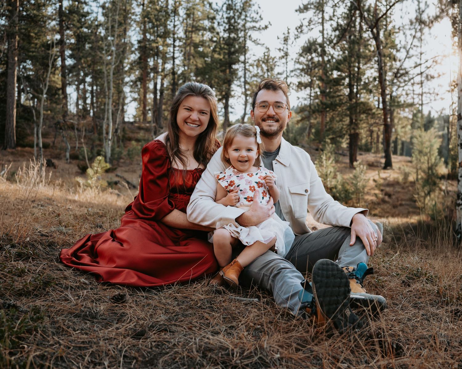 Family relaxing together on forest floor surrounded by tall pine trees.
