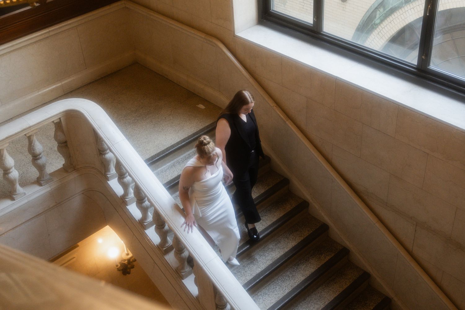 A wedding couple walks up a bright indoor staircase with natural light streaming through windows.