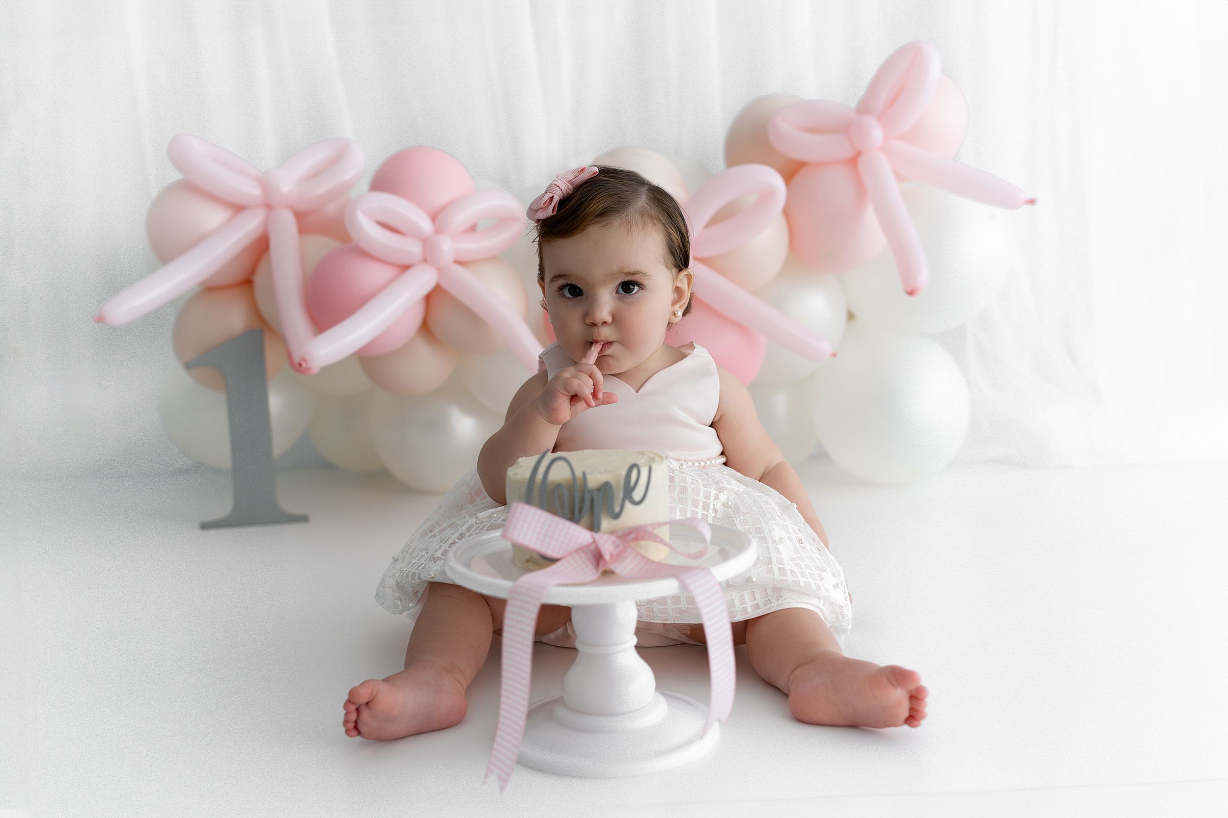 Baby sits on white chair surrounded by pink bow decorations and balloons.