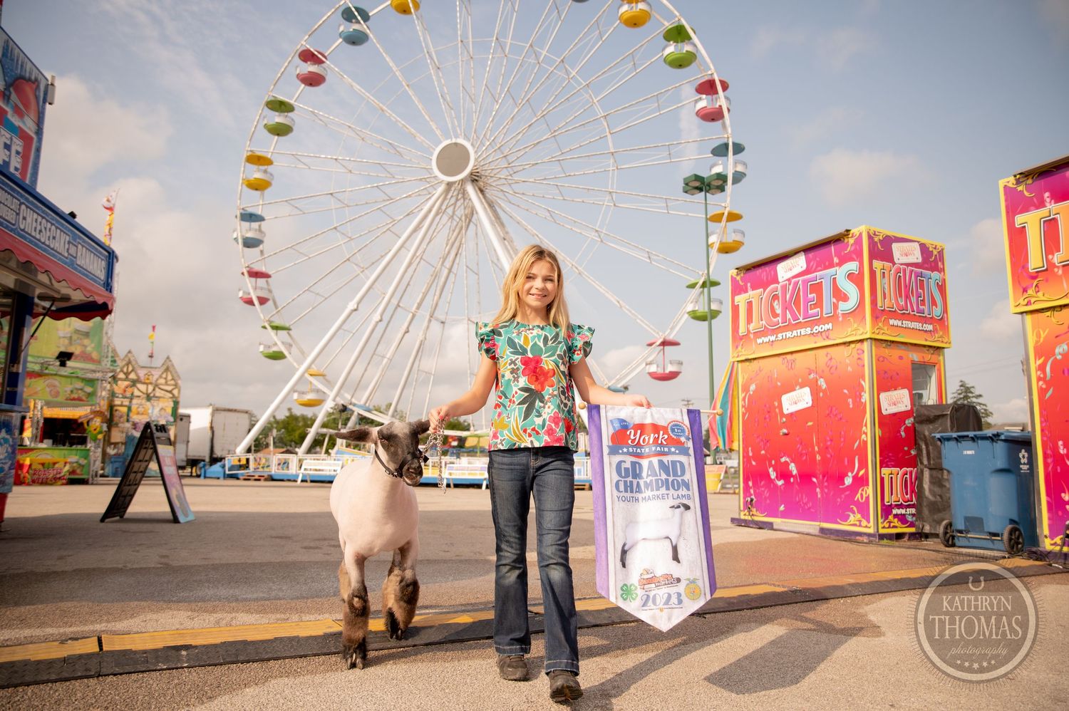 A person walks a large animal past a colorful ferris wheel and carnival booths at a state fair.