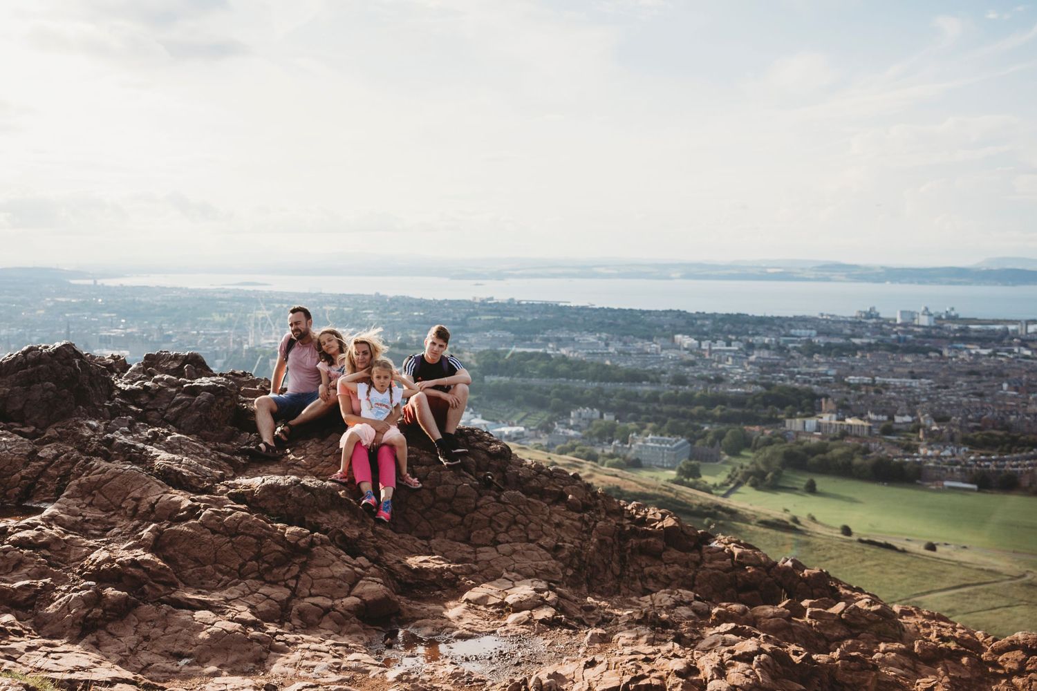 Group sitting on rocky outcrop overlooking scenic city and water views.