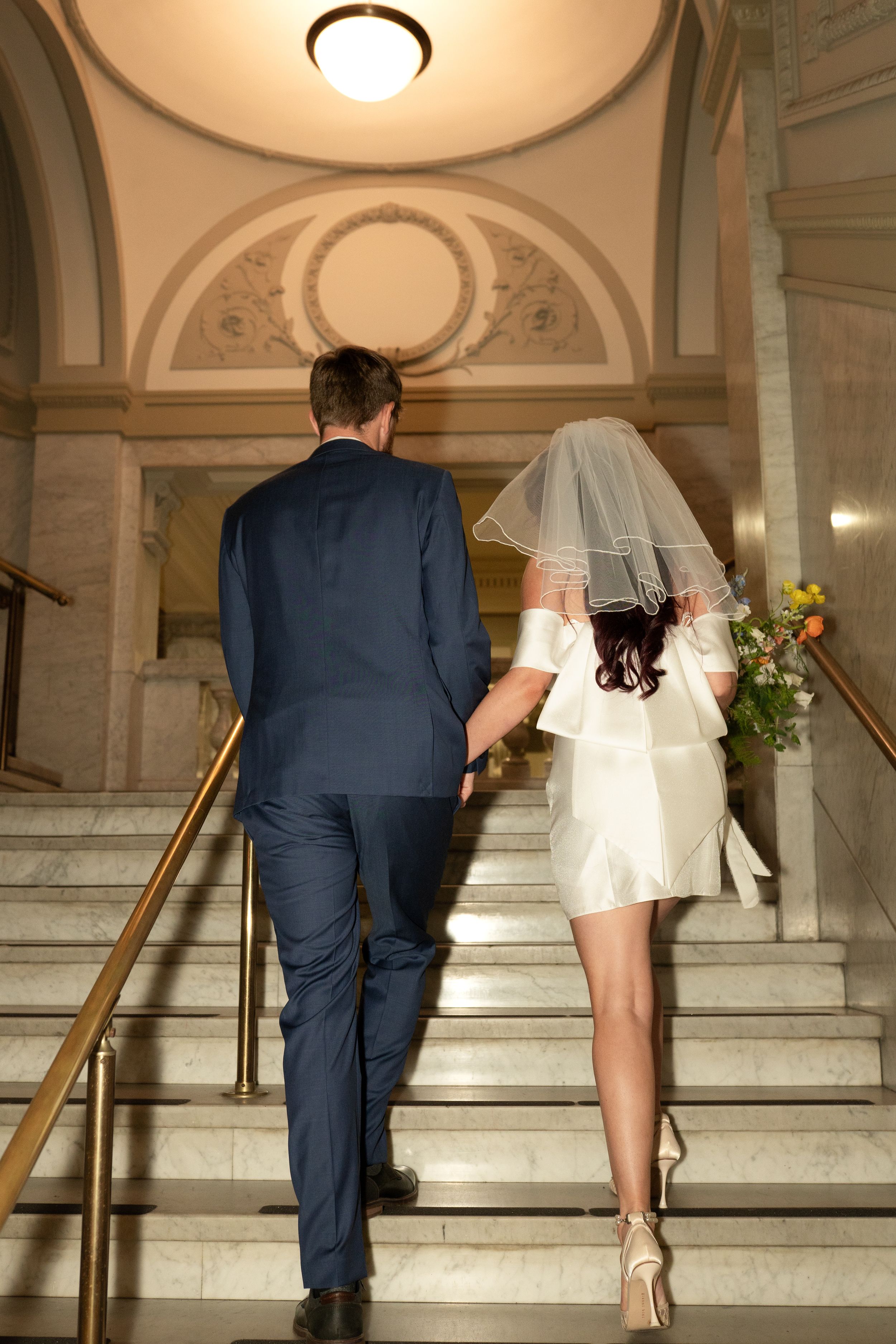 Wedding couple ascends marble staircase in grand historic building.