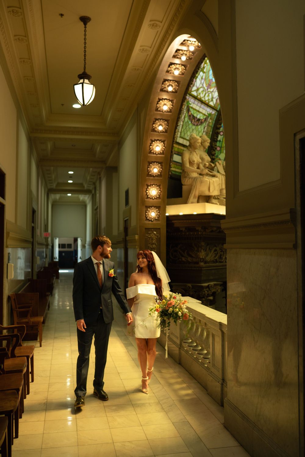 Couple walking through ornate hallway with arched windows and marble columns.