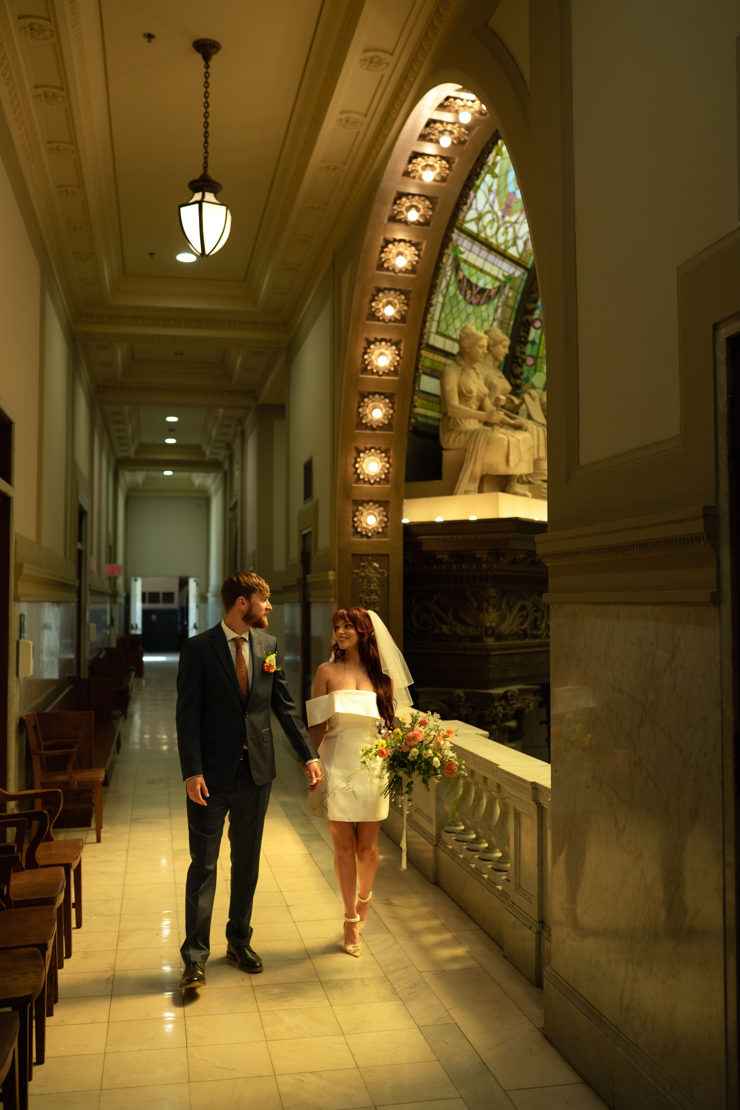 Couple walking through ornate hallway with arched windows and marble columns.