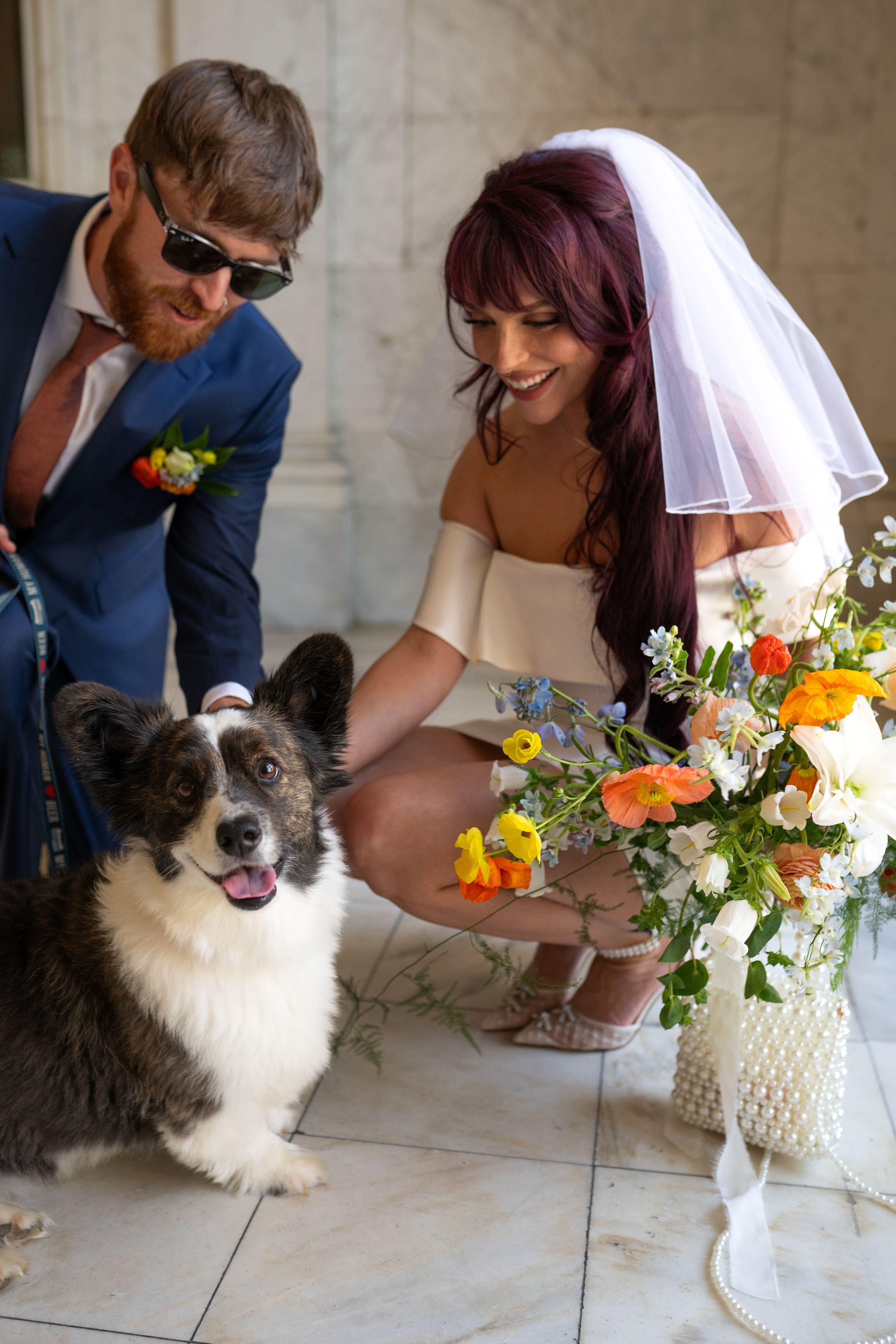 Wedding couple interacting with fluffy dog next to floral arrangement.