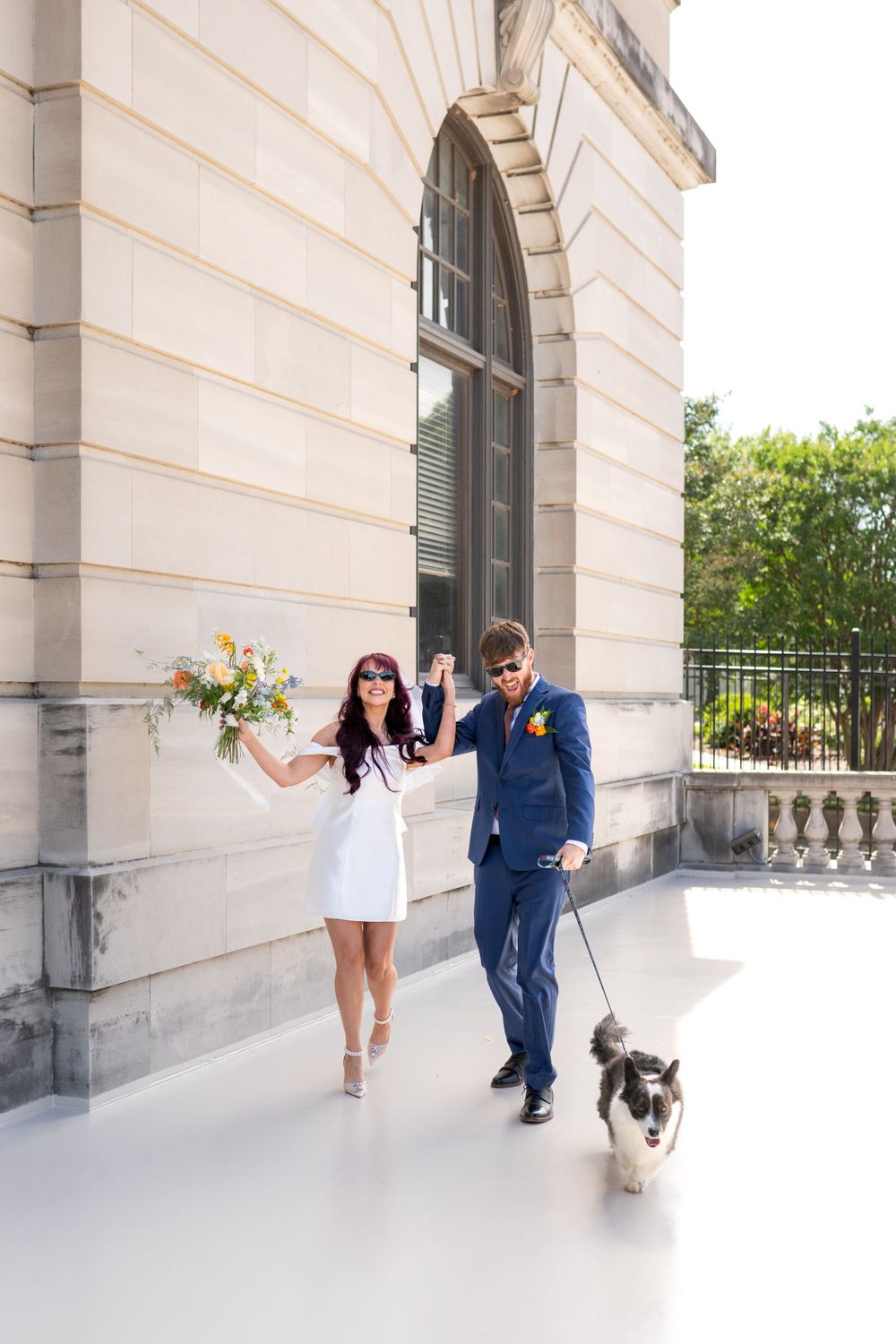 A couple walks along a elegant columned walkway with a small dog as the bride holds a bright floral bouquet.